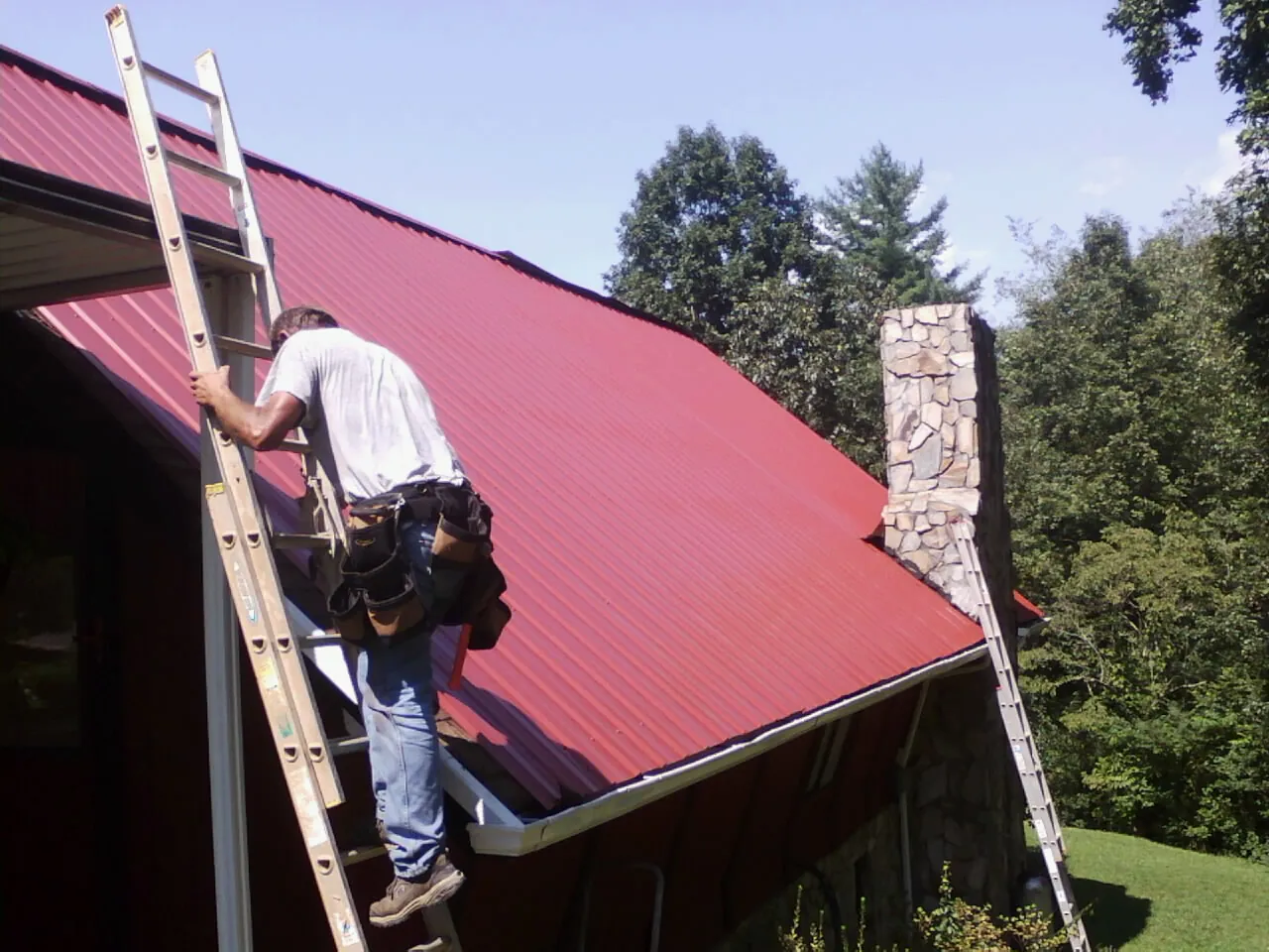 Roofer on a ladder beside a red metal roof and stone chimney — Soffit & Fascia Repair by Roofing Brothers in Hackleburg